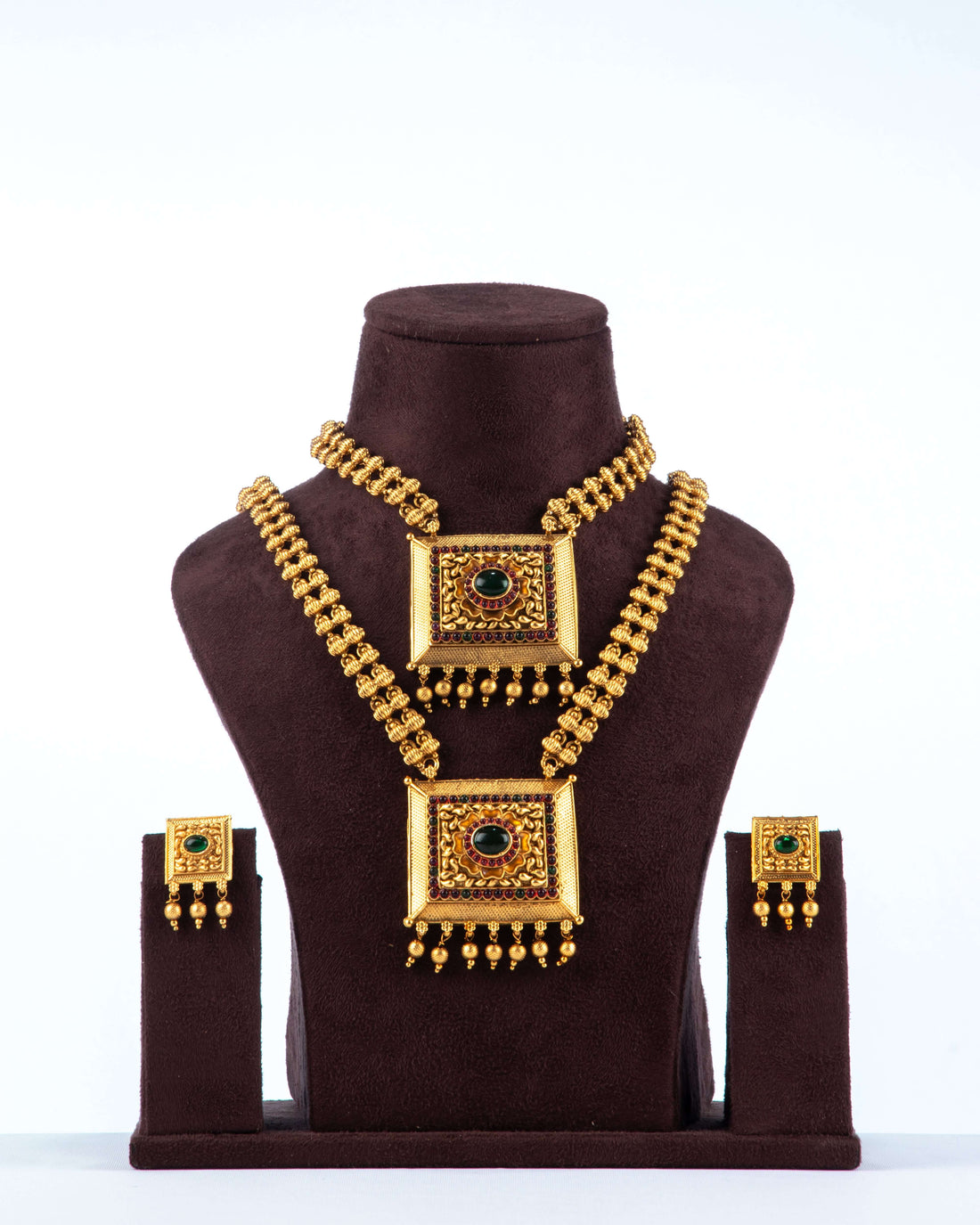 A traditional South Indian dual necklace set with square pendants, featuring a short and a long chain, displayed with matching earrings on a brown jewelry bust. The gold-toned set has intricate designs and is adorned with red and green stones.