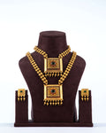 A traditional South Indian dual necklace set with square pendants, featuring a short and a long chain, displayed with matching earrings on a brown jewelry bust. The gold-toned set has intricate designs and is adorned with red and green stones.