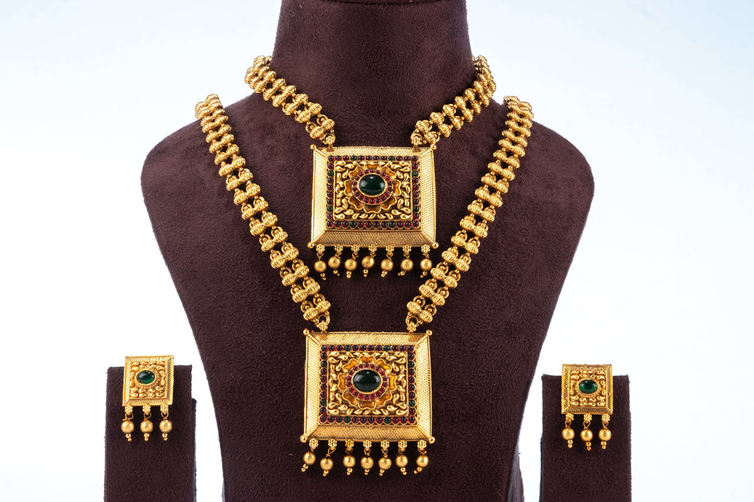A traditional South Indian dual necklace set with square pendants, featuring a short and a long chain, displayed with matching earrings on a brown jewelry bust. The gold-toned set has intricate designs and is adorned with red and green stones.