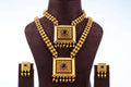 A traditional South Indian dual necklace set with square pendants, featuring a short and a long chain, displayed with matching earrings on a brown jewelry bust. The gold-toned set has intricate designs and is adorned with red and green stones.