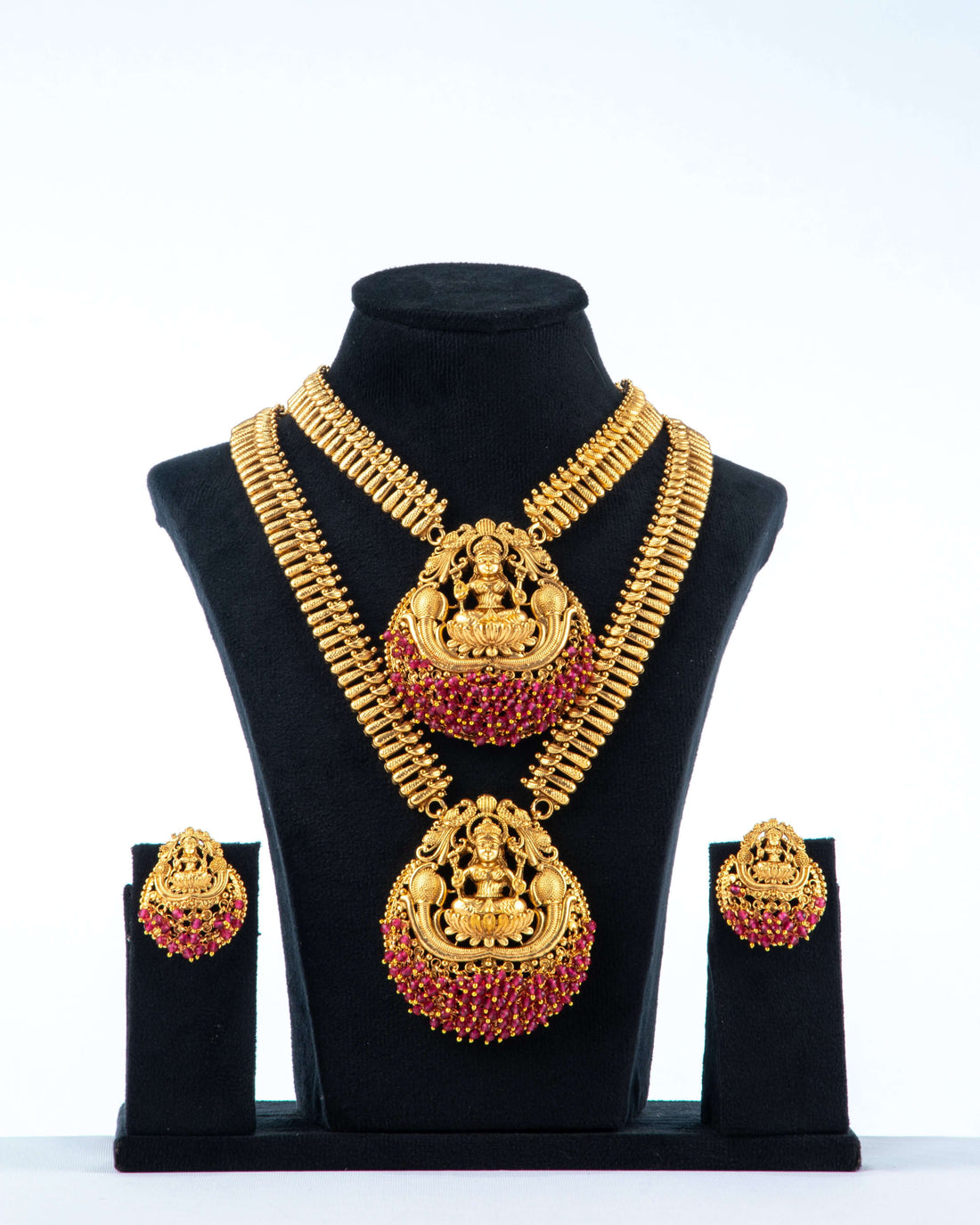 A traditional long temple necklace set with Laxmi pendants and matching earrings. The set features two gold-toned necklaces with intricate chains, both adorned with large, round pendants that depict the goddess Laxmi. The pendants are decorated with ruby-red stones and have a red beaded fringe. The matching earrings are also round with a Laxmi motif and red stone accents. The jewelry is displayed on a black bust against a white background.