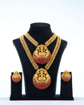 A traditional long temple necklace set with Laxmi pendants and matching earrings. The set features two gold-toned necklaces with intricate chains, both adorned with large, round pendants that depict the goddess Laxmi. The pendants are decorated with ruby-red stones and have a red beaded fringe. The matching earrings are also round with a Laxmi motif and red stone accents. The jewelry is displayed on a black bust against a white background.