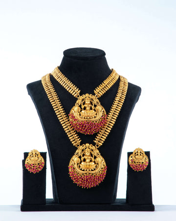 A traditional long temple necklace set with Laxmi pendants and matching earrings. The set features two gold-toned necklaces with intricate chains, both adorned with large, round pendants that depict the goddess Laxmi. The pendants are decorated with ruby-red stones and have a red beaded fringe. The matching earrings are also round with a Laxmi motif and red stone accents. The jewelry is displayed on a black bust against a white background.
