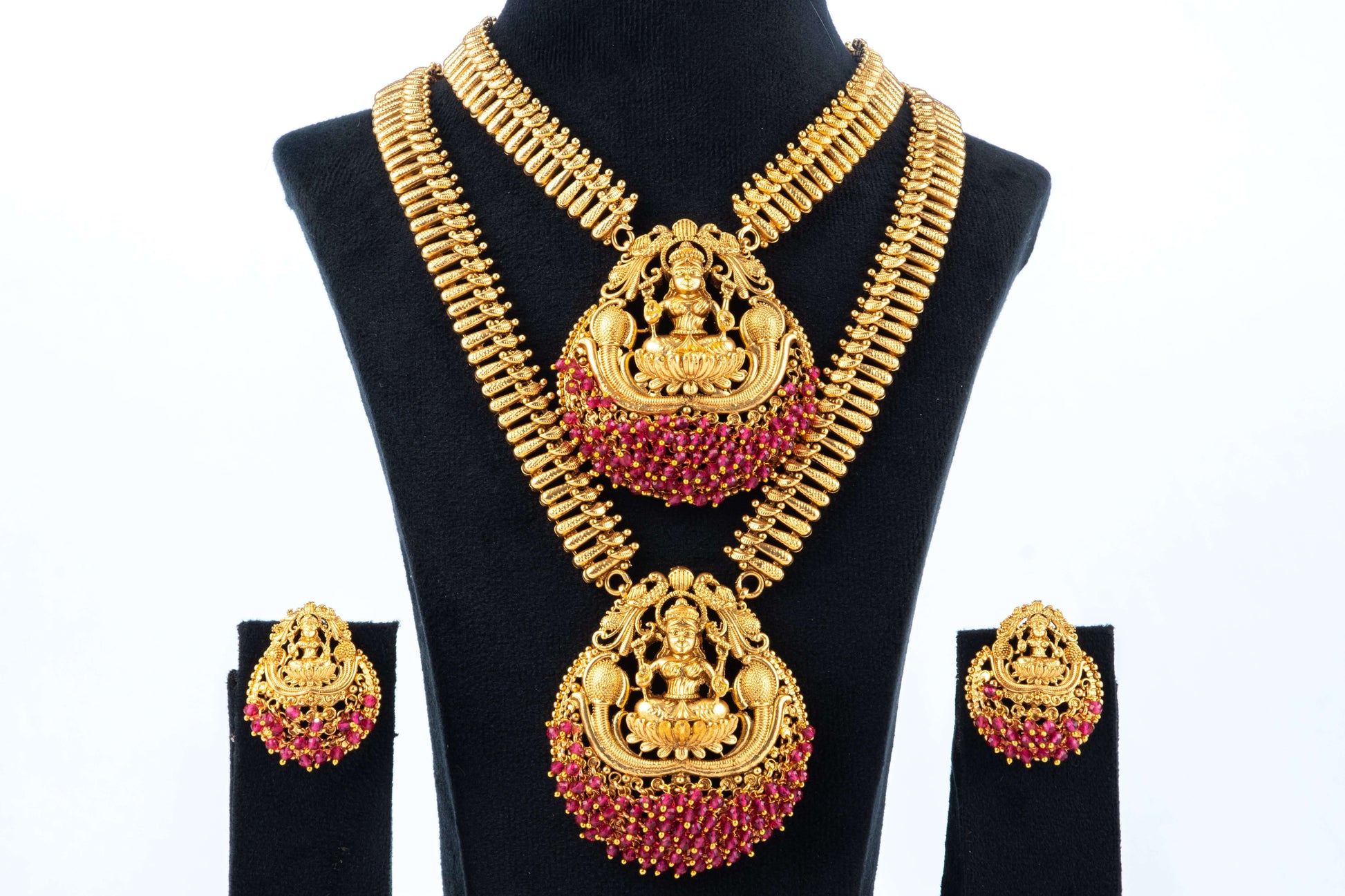 A Traditional Long Temple Necklace set with Laxmi Pendants is shown, featuring a gold-toned two-piece necklace with intricate chains and matching earrings. Each piece has a large, central pendant depicting the goddess Laxmi surrounded by ruby-red beads. The jewelry is displayed on a black mannequin bust.