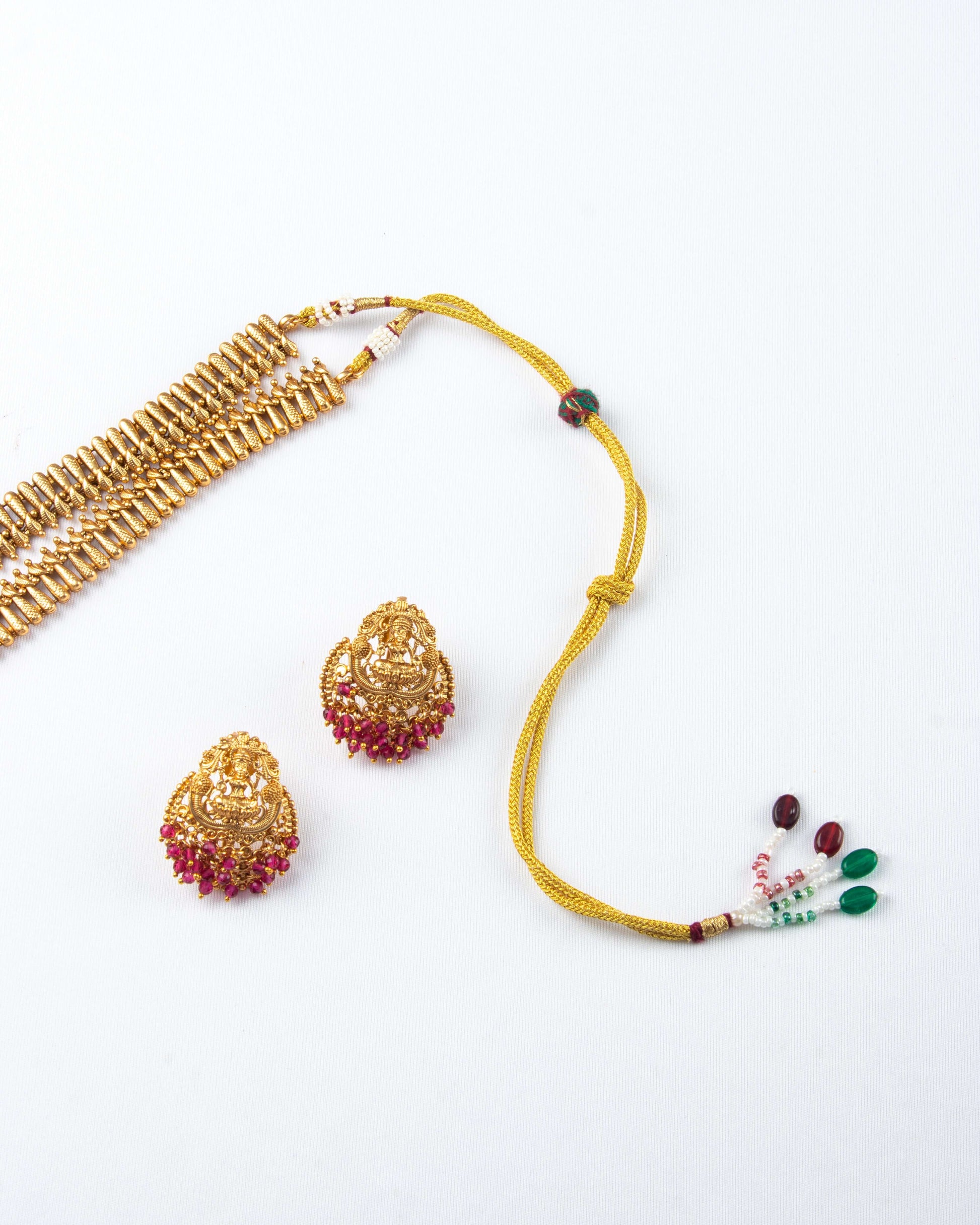 Laxmi pendants is displayed on a white surface. The set includes a gold-toned, multi-strand necklace and matching earrings, all featuring a prominent Laxmi pendant and detailed red bead accents. The necklace has a beaded adjustable string on the end.