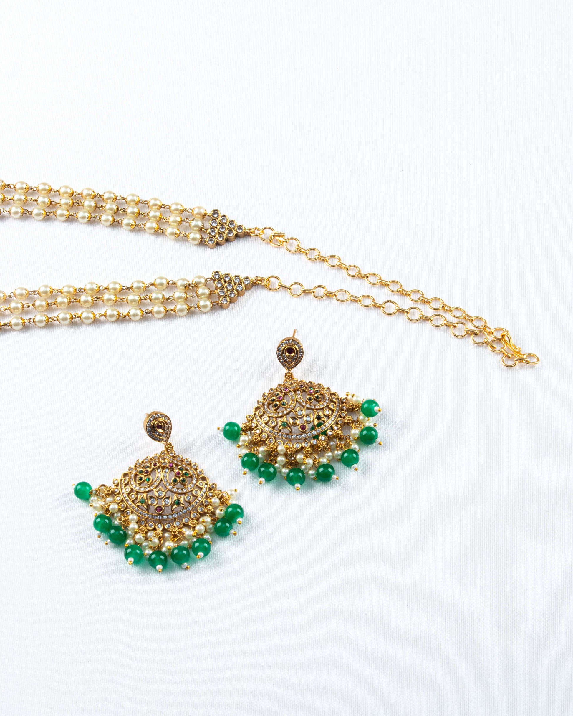A White Pearl Cascade Necklace set with Green Pearl Pendant is displayed on a white surface. The set includes a multi-strand necklace of white pearls connected by a gold-toned, ornate clasp. A pair of matching, intricate gold earrings with red and green stones and a border of dangling green pearl drops are shown in the foreground.