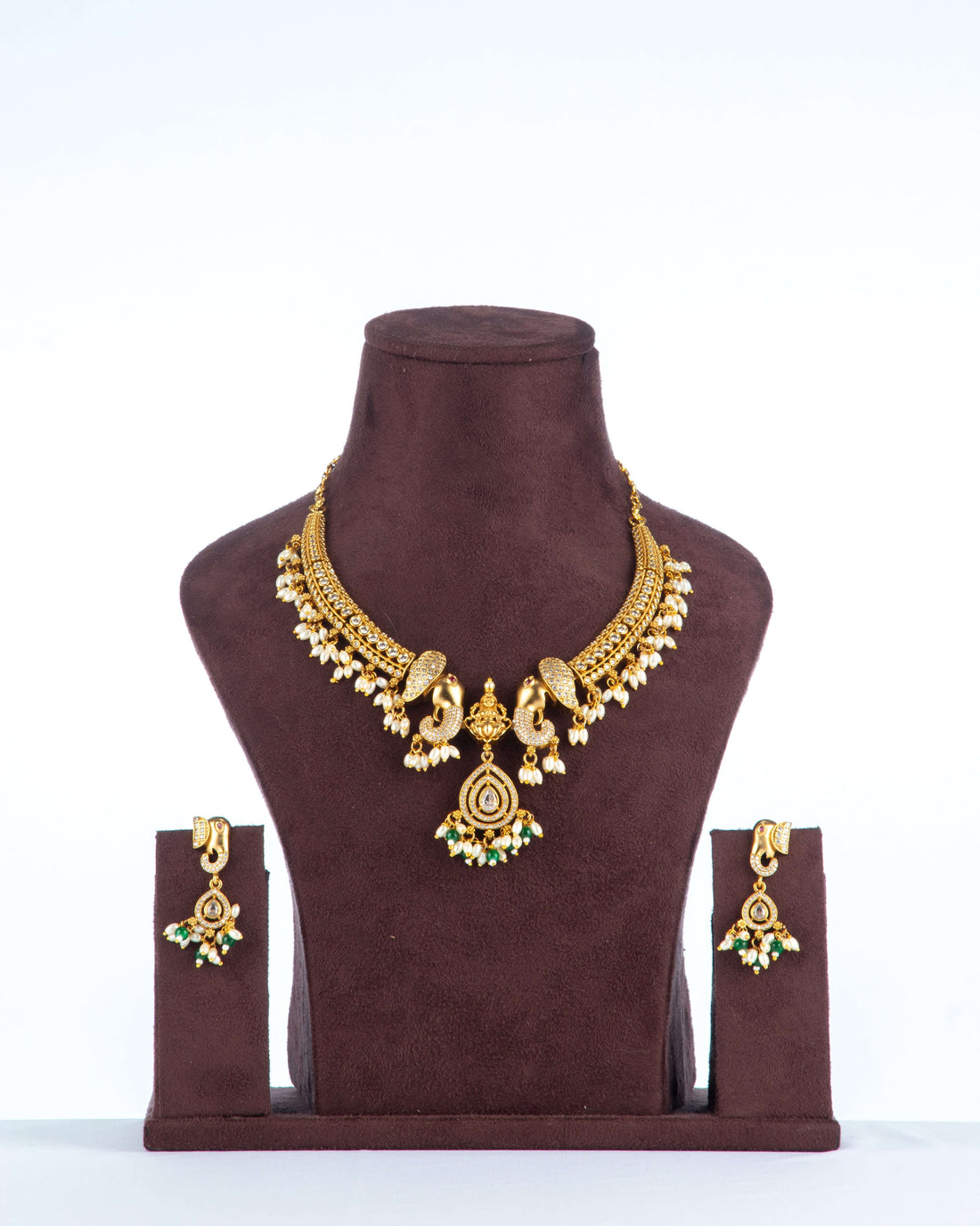 A traditional Gold Temple Jewellery Necklace and earring set is displayed on a brown velvet bust against a white background. The necklace features a broad, antique gold-toned design with intricate peacock motifs, adorned with pearls and green stone accents, and a central drop pendant with a green stone and pearl cluster. The matching stud earrings have a similar peacock design with a single green stone.
