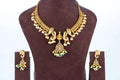 A traditional Gold Temple Jewellery Necklace set is displayed on a brown velvet bust. The necklace features a broad, antique gold-toned choker design with intricate peacock motifs, adorned with rows of white pearls and a central teardrop pendant with green and white beads. The matching stud earrings have a similar peacock design and dangle with clusters of green and white beads.