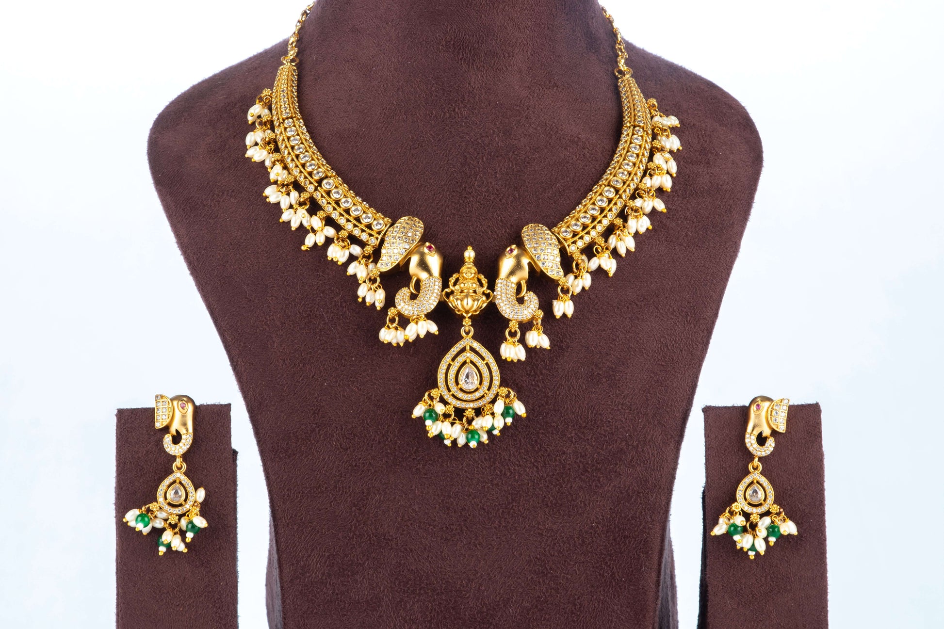 A traditional Gold Temple Jewellery Necklace set is displayed on a brown velvet bust. The necklace features a broad, antique gold-toned choker design with intricate peacock motifs, adorned with rows of white pearls and a central teardrop pendant with green and white beads. The matching stud earrings have a similar peacock design and dangle with clusters of green and white beads.