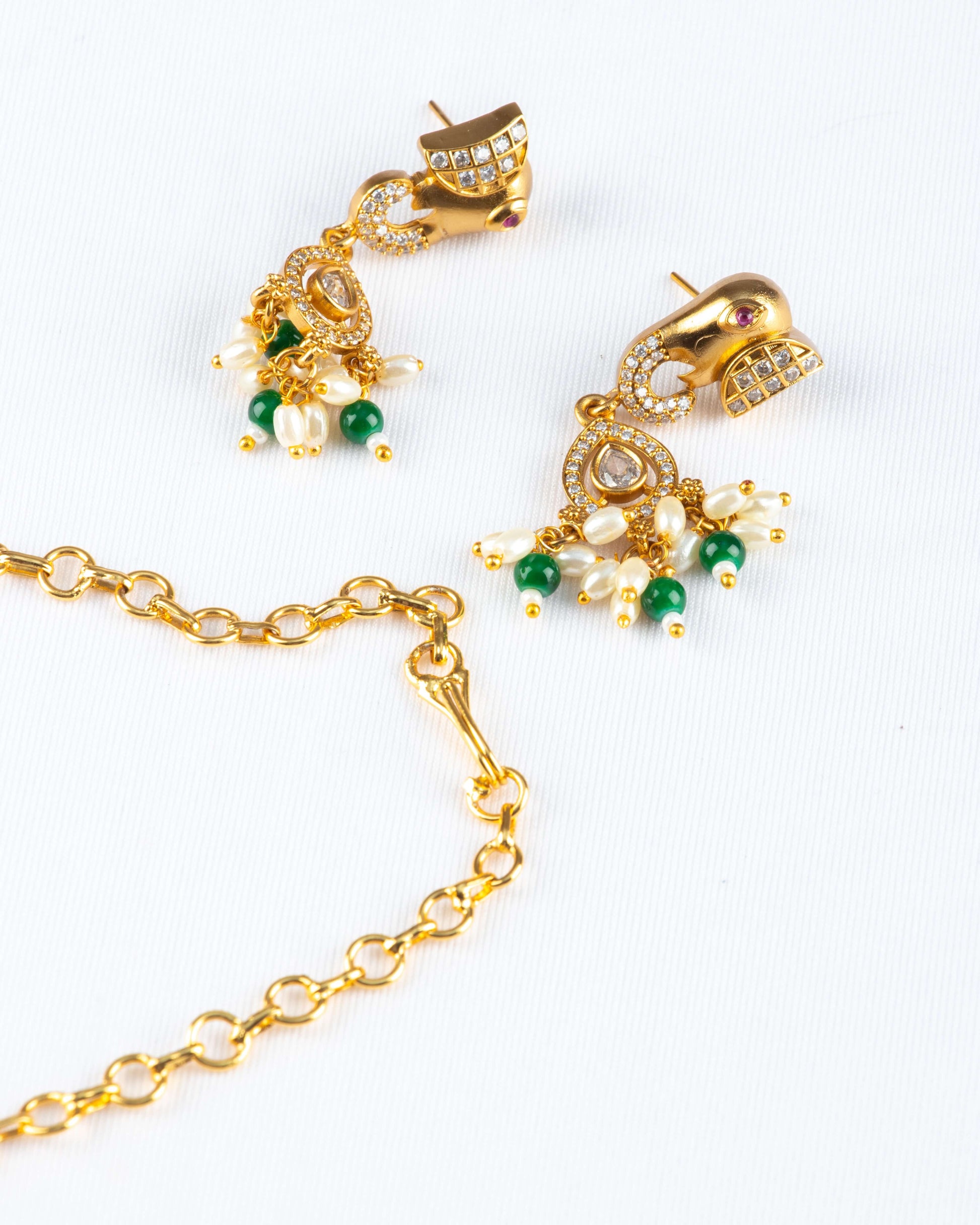 A pair of intricate earrings and a section of a chain from a Gold Temple Jewellery Necklace are displayed on a white background. The gold-toned earrings feature a stylized peacock or elephant head design, embellished with clear and red stones. Dangling from the earrings are clusters of white pearls and green beads. A simple gold chain is visible in the foreground.