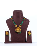 A beaded long necklace set with green beads and a large, intricate gold pendant is displayed on a brown jewelry bust. The set includes a pair of matching gold earrings with small green beads hanging from them.