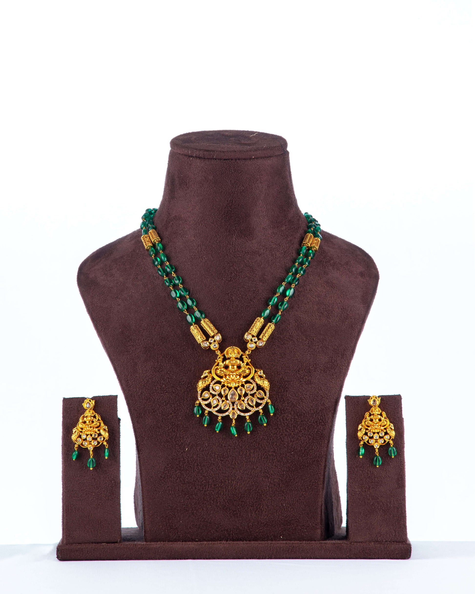 A beaded long necklace set with green beads and a large, intricate gold pendant is displayed on a brown jewelry bust. The set includes a pair of matching gold earrings with small green beads hanging from them.