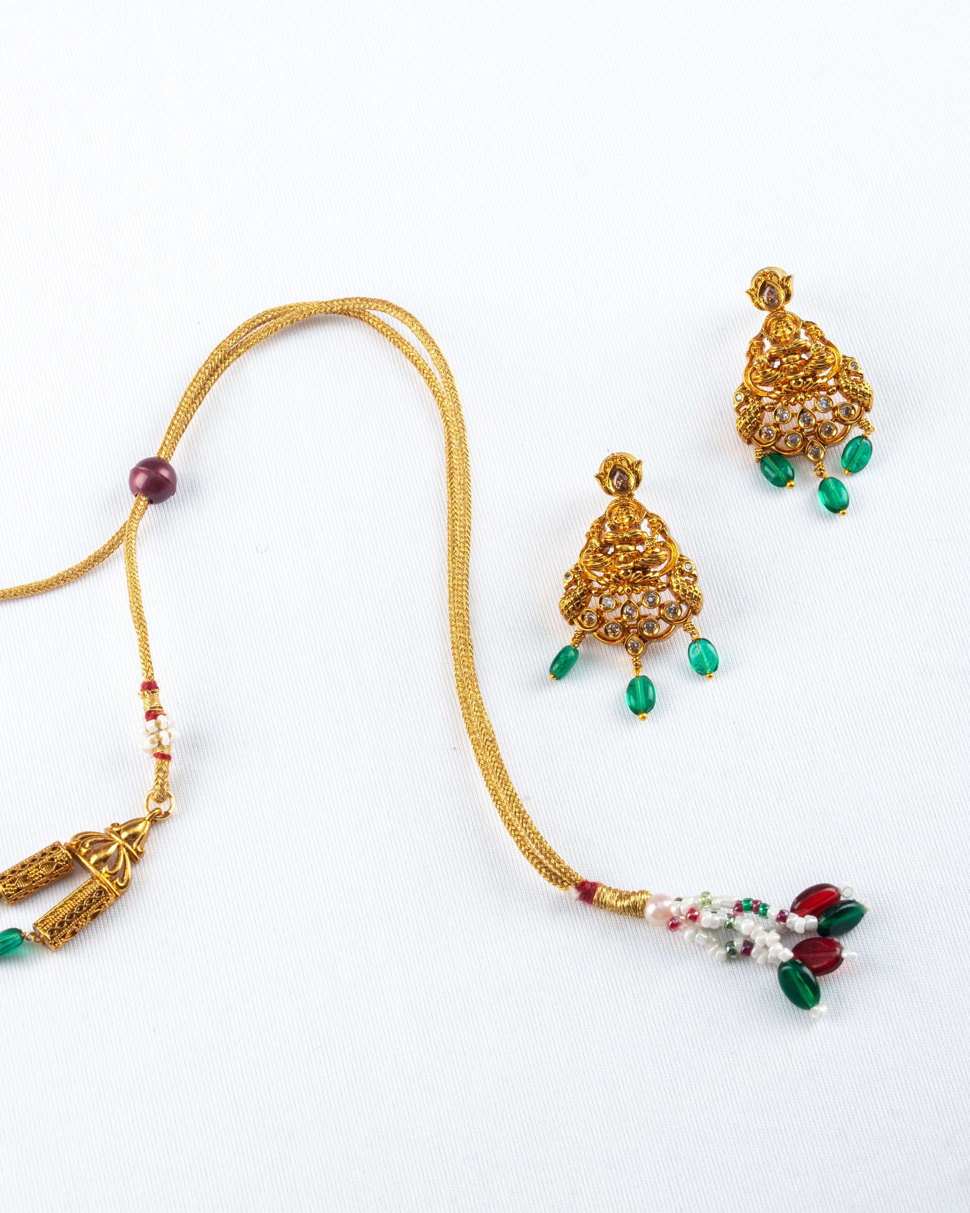 beaded long necklace set on a white background. The set includes a pair of intricate, gold-toned jhumka earrings with clear stones and green bead drops, along with a portion of the adjustable necklace cord. The necklace cord has decorative gold, red, green, and white beads at the end and a single red adjustable bead.