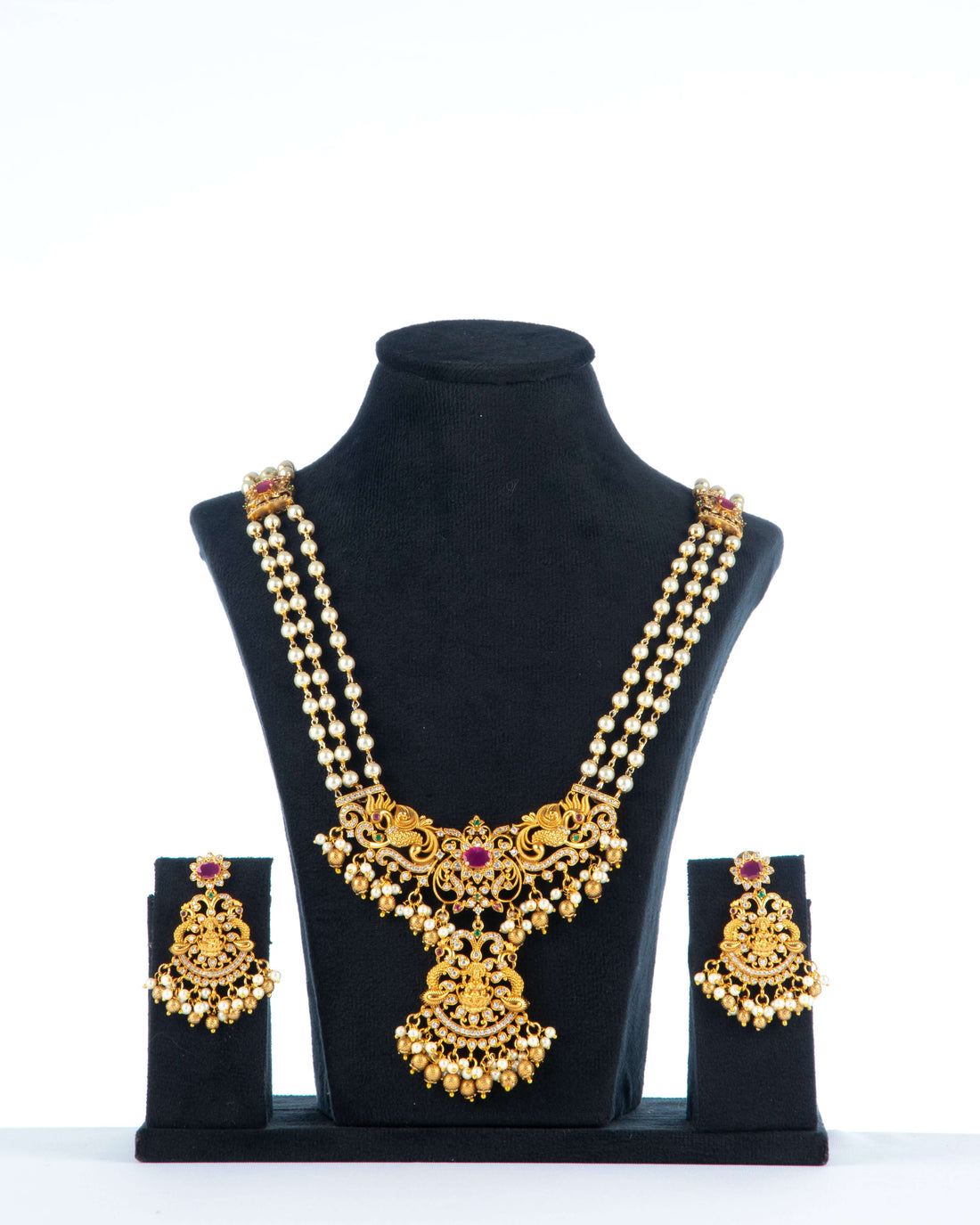 A long, multi-strand beaded necklace set with matching earrings is displayed on a black jewelry bust. The gold-toned antique necklace features a prominent pendant with intricate detailing, a central ruby-red stone, and pearl drop accents. The matching earrings have a similar design with a red stone and pearl drops.