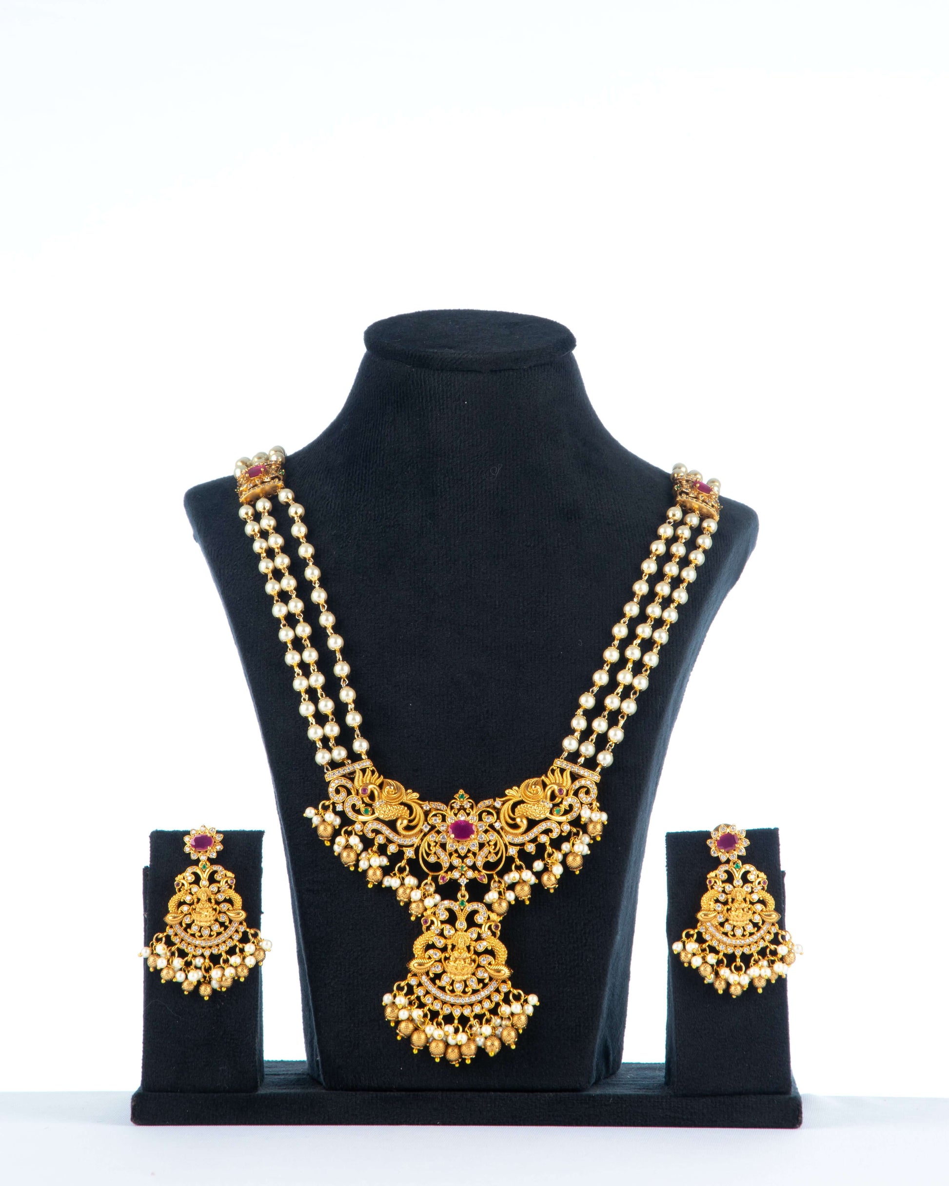 A long, multi-strand beaded necklace set with matching earrings is displayed on a black jewelry bust. The gold-toned antique necklace features a prominent pendant with intricate detailing, a central ruby-red stone, and pearl drop accents. The matching earrings have a similar design with a red stone and pearl drops.