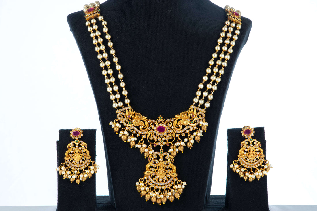 A long, multi-strand beaded necklace set with matching earrings is displayed on a black jewelry bust. The gold-toned antique necklace features a prominent pendant with intricate detailing, a central ruby-red stone, and pearl drop accents. The matching earrings have a similar design with a red stone and pearl drops.