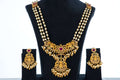 A long, gold-toned antique beaded necklace set with matching earrings is displayed on a black jewelry bust. The necklace features multiple strands of white beads and a prominent, ornate pendant with a central red stone and pearl drop accents. The matching earrings have a similar design with red stones and pearl drops.