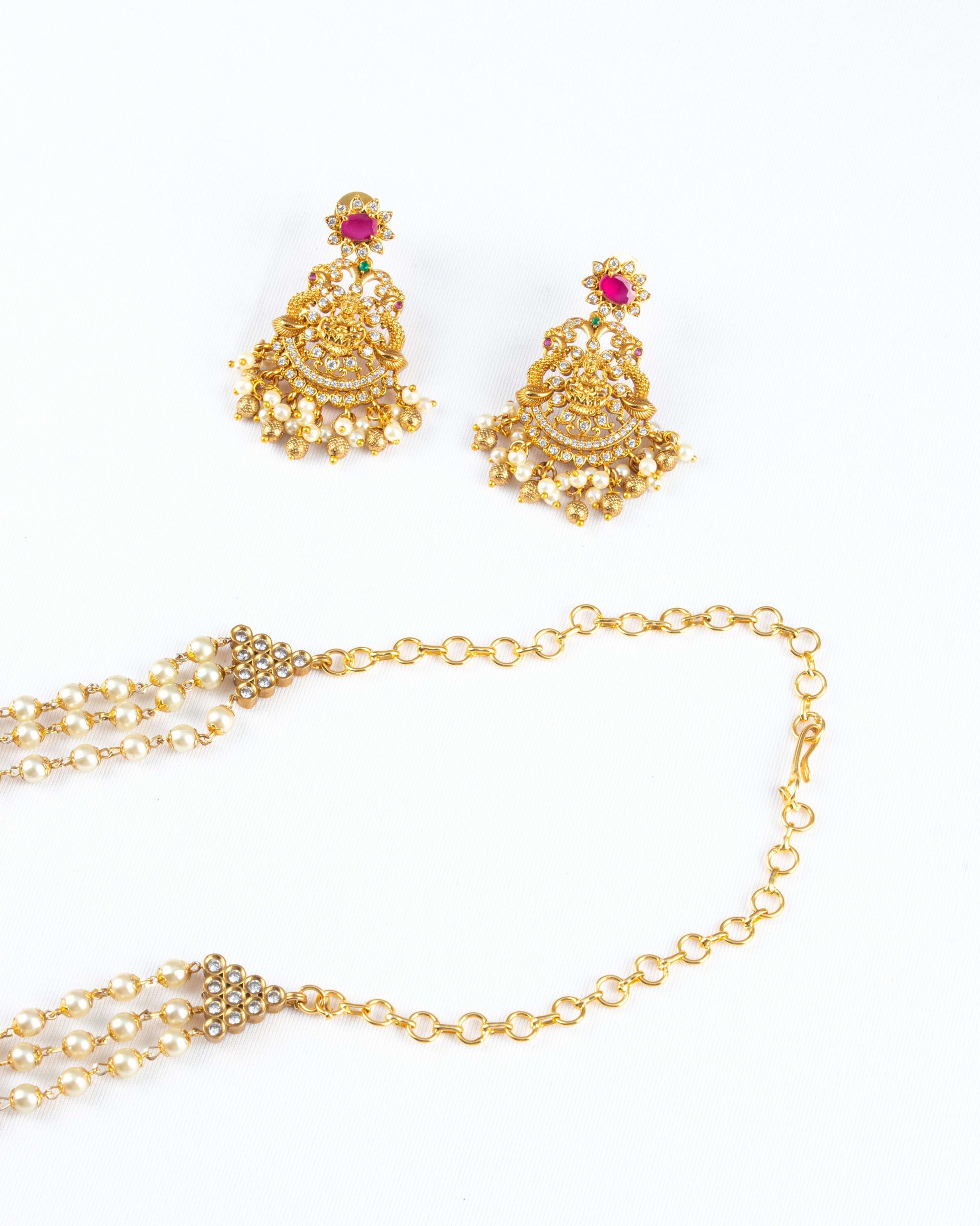 A long, gold-toned antique beaded necklace set with matching earrings is displayed on a white surface. The necklace features multiple strands of pearls and a decorative clasp, while the intricate, dangling earrings have a large ruby-red stone and a fringe of pearls.