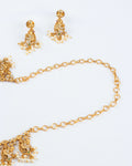 A gold-toned necklace and earring set is arranged on a white surface. The necklace features a simple chain connected to an ornate pendant decorated with small white stones, a green stone, and hanging pearls. The matching earrings have a similar design with a stud and dangling pearls.