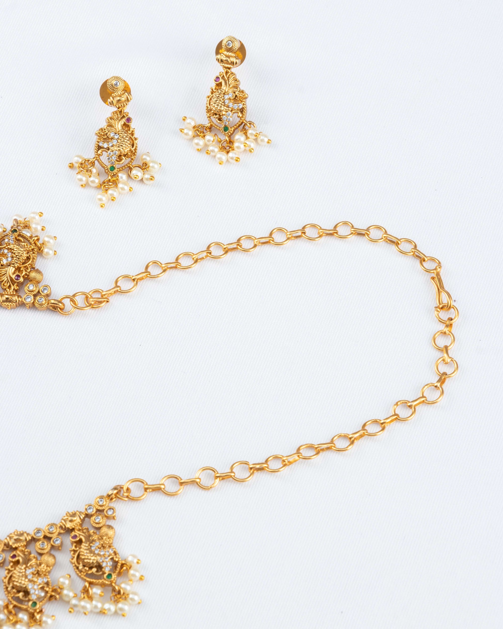 A gold-toned necklace and earring set is arranged on a white surface. The necklace features a simple chain connected to an ornate pendant decorated with small white stones, a green stone, and hanging pearls. The matching earrings have a similar design with a stud and dangling pearls.