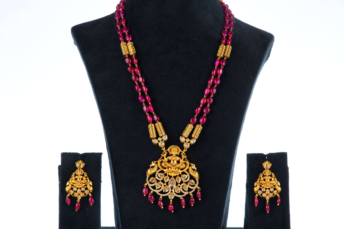 A beaded long necklace set with intricate gold-colored temple jewelry pendant and coordinating earrings, displayed on a mannequin bust.