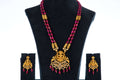 A gold and red beaded long necklace set with a large, intricate pendant featuring a central figure surrounded by stones and dangling red beads. The set includes a pair of matching earrings, and all pieces are displayed on black velvet busts against a white background.
