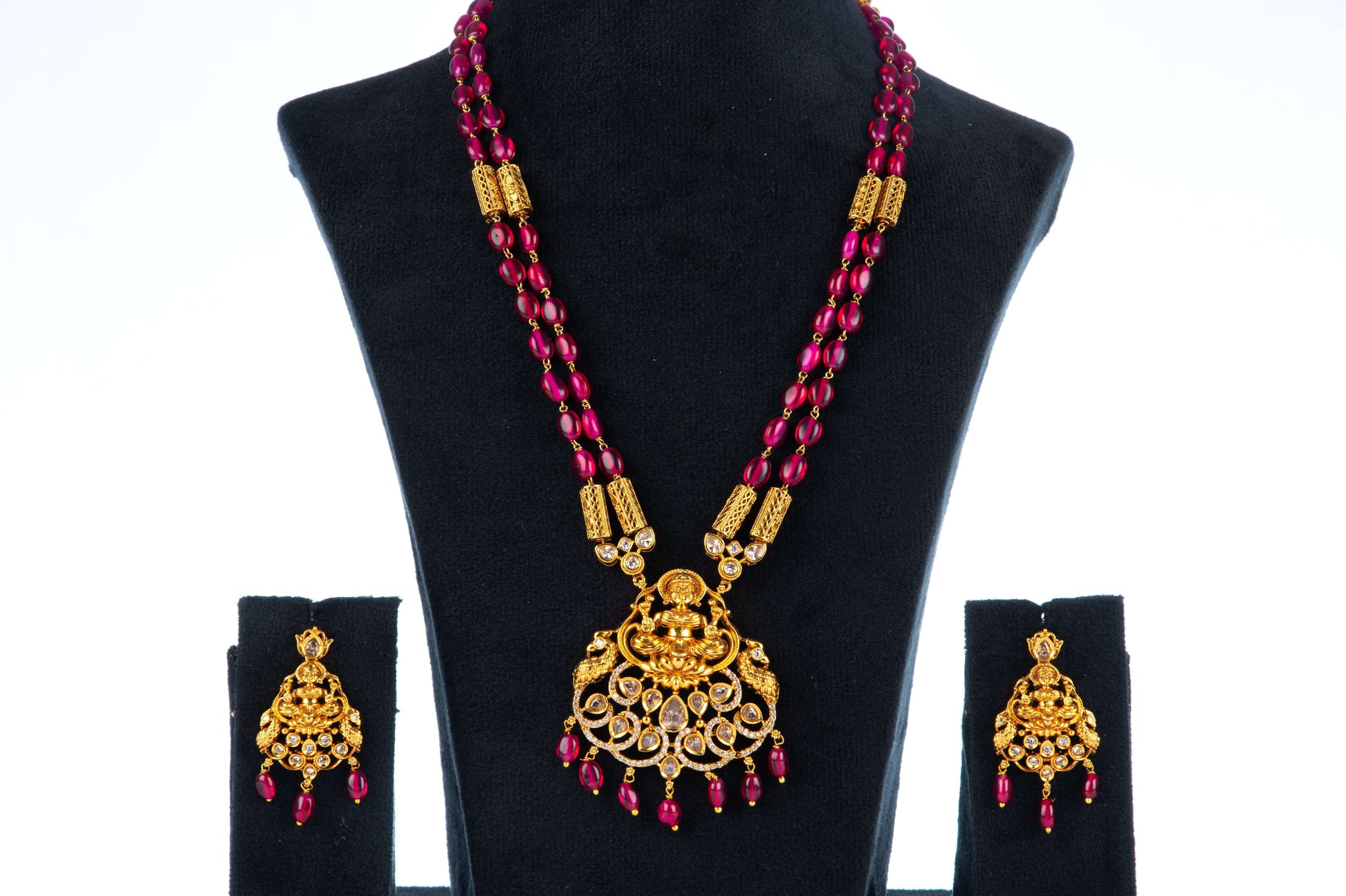 A gold and red beaded long necklace set with a large, intricate pendant featuring a central figure surrounded by stones and dangling red beads. The set includes a pair of matching earrings, and all pieces are displayed on black velvet busts against a white background.