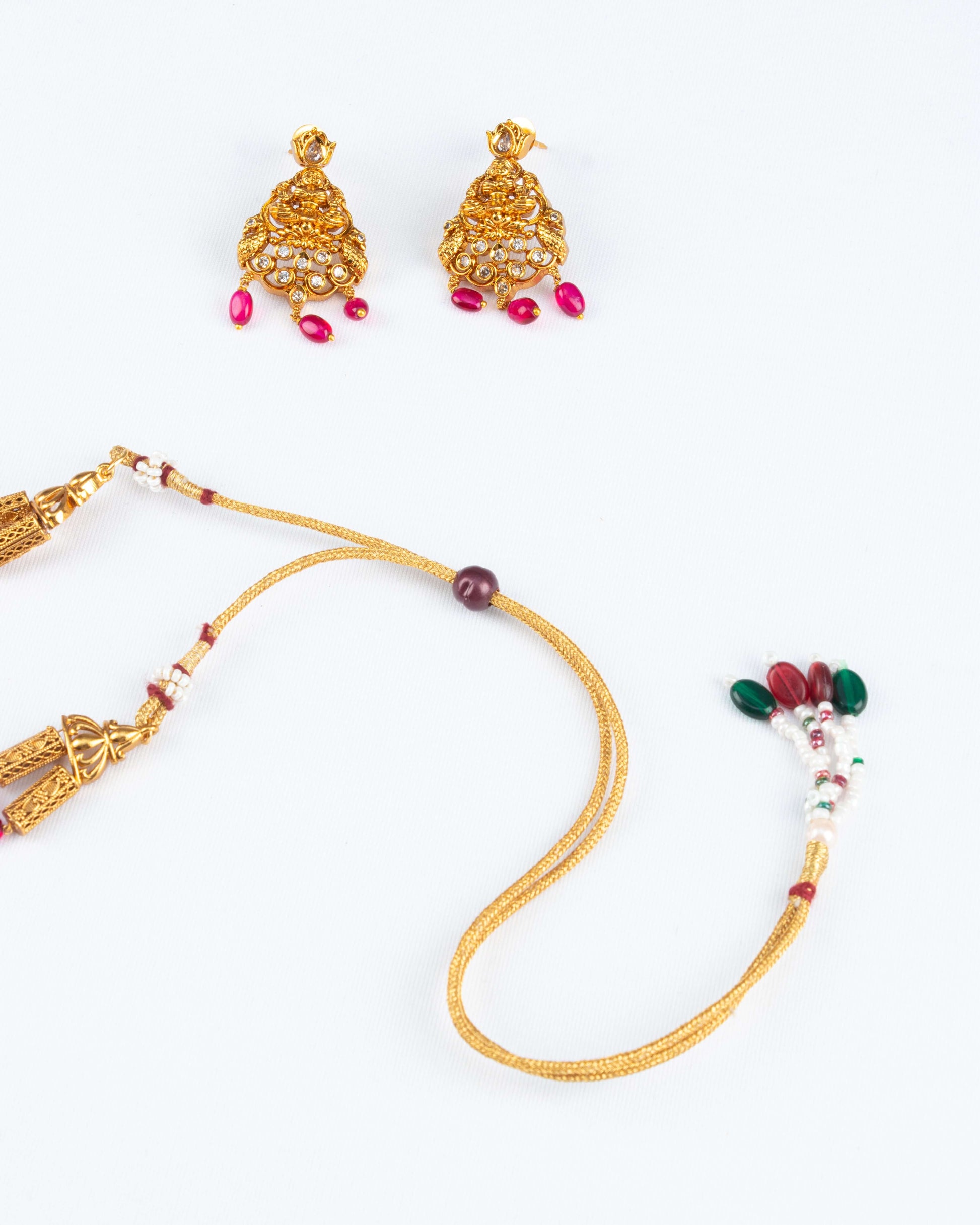 Pair of traditional gold earrings and the adjustable back cord of a matching necklace, both set against a white background. The earrings are intricate, featuring a bell-shaped design with clear stones and small, dangling pink or red beads. The necklace cord is gold-colored with a sliding red bead for adjustment and a decorative end piece with red, green, and white beads.