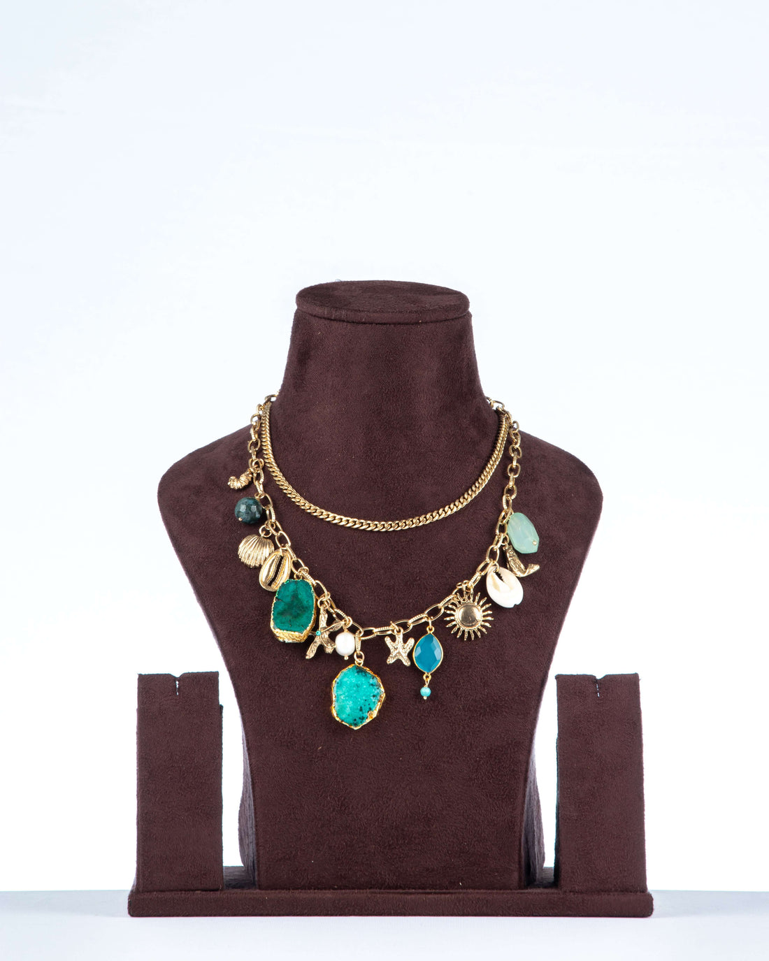 A double-layered chunky agate necklace, featuring two large, irregularly shaped teal-colored agate pendants as the focal points. The gold-toned chain is adorned with various charms, including seashells, starfish, and a sun, giving it a beach-inspired aesthetic. The necklace is displayed on a brown jewelry bust.
