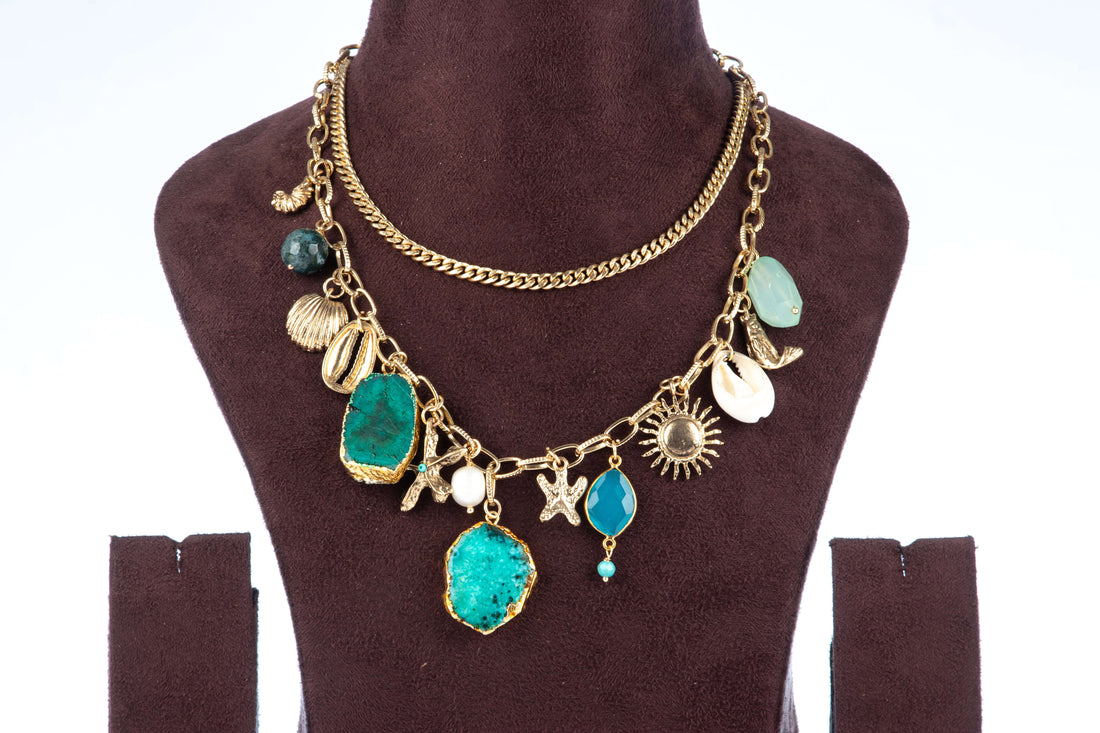 A double-layered chunky agate necklace, featuring two large, irregularly shaped teal-colored agate pendants as the focal points. The gold-toned chain is adorned with various charms, including seashells, starfish, and a sun, giving it a beach-inspired aesthetic. The necklace is displayed on a brown jewelry bust.