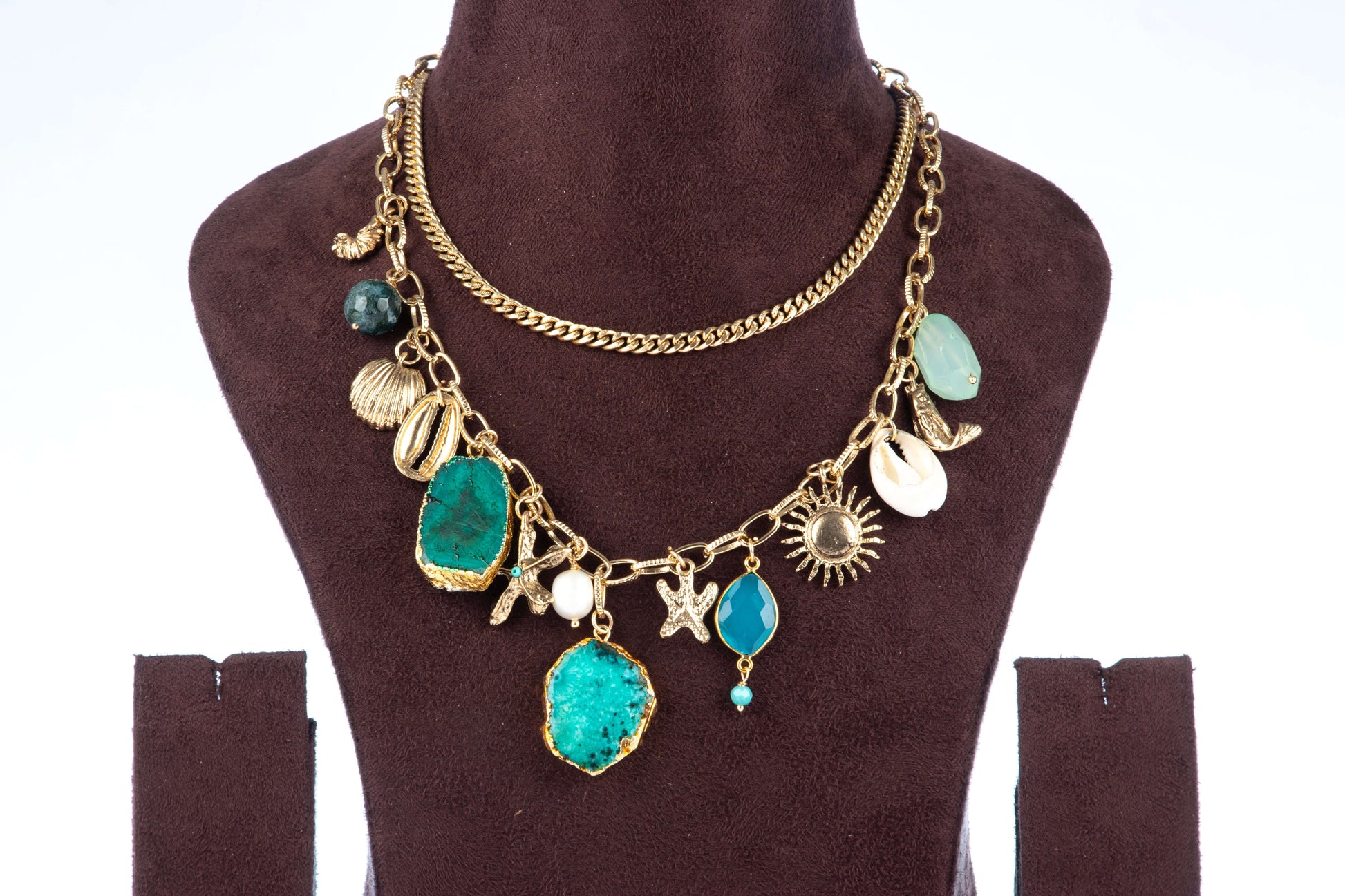 A double-layered chunky agate necklace, featuring two large, irregularly shaped teal-colored agate pendants as the focal points. The gold-toned chain is adorned with various charms, including seashells, starfish, and a sun, giving it a beach-inspired aesthetic. The necklace is displayed on a brown jewelry bust.
