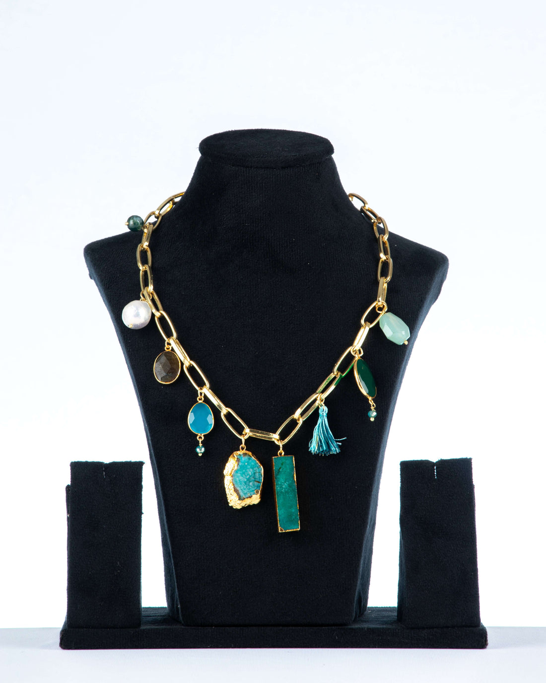 A single-layered, chunky Agate necklace with a gold-toned chain is displayed on a black jewelry bust. The necklace features a variety of charms and pendants, including large, irregularly shaped blue and green agate stones, smaller round stones, a pearl, and a blue tassel.