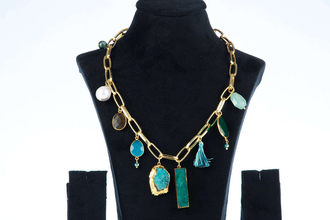 A single-layered, chunky Agate necklace with a gold-toned chain is displayed on a black jewelry bust. The necklace features a variety of charms and pendants, including large, irregularly shaped blue and green agate stones, smaller round stones, a pearl, and a blue tassel.
