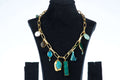 A single-layered, chunky Agate necklace with a gold-toned chain is displayed on a black jewelry bust. The necklace features a variety of charms and pendants, including large, irregularly shaped blue and green agate stones, smaller round stones, a pearl, and a blue tassel.