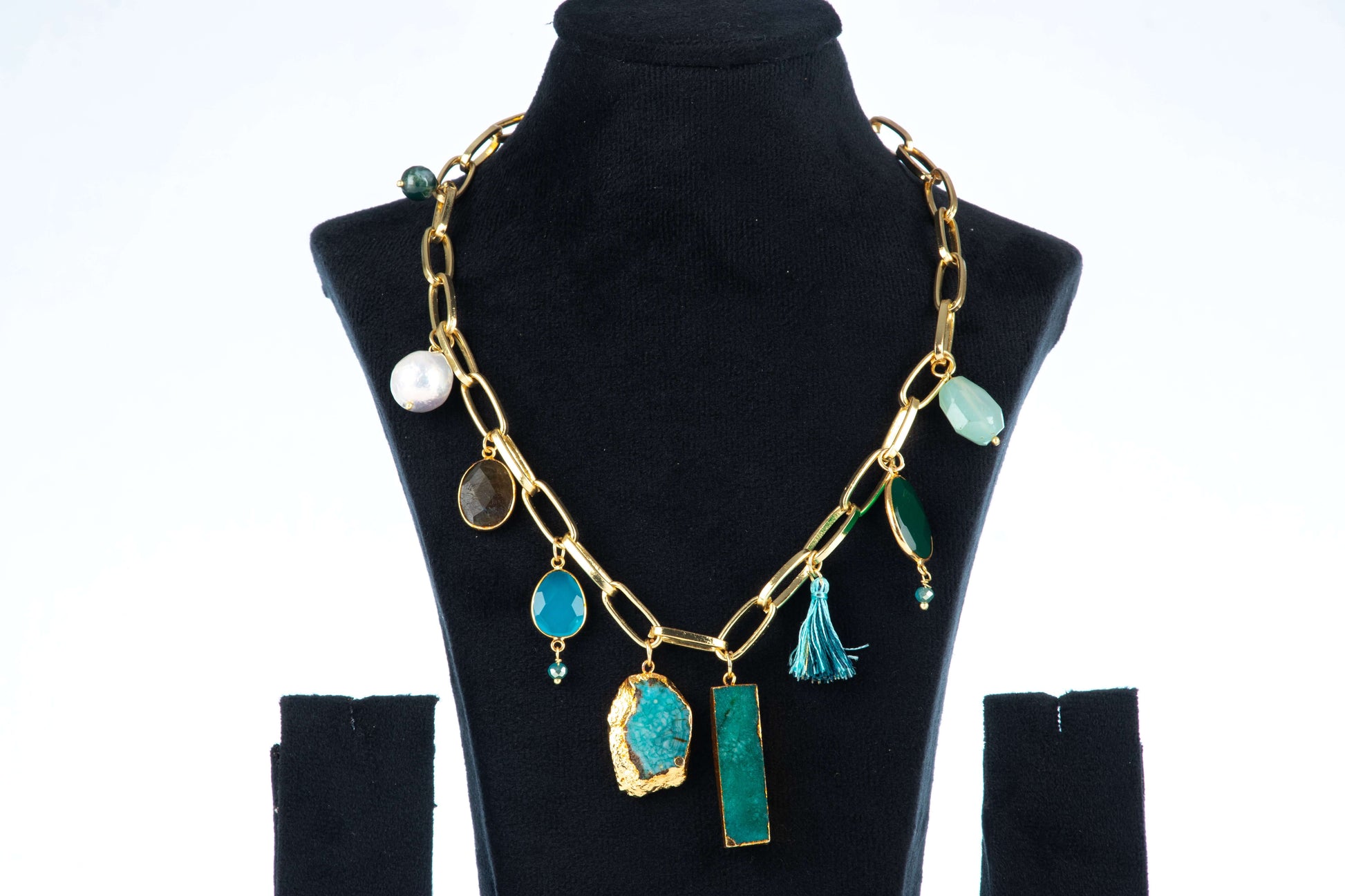 A single-layered, chunky Agate necklace with a gold-toned chain is displayed on a black jewelry bust. The necklace features a variety of charms and pendants, including large, irregularly shaped blue and green agate stones, smaller round stones, a pearl, and a blue tassel.