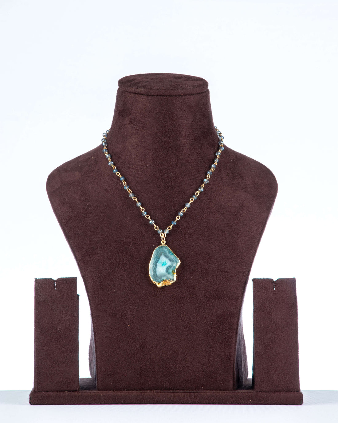 A Full beaded agate pendant necklace is shown on a brown velvet jewelry bust against a light background. The necklace features a gold-toned chain with small, faceted green beads and a large, gold-edged agate pendant with a blue-green, crystalline center.
