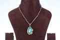 A Full beaded agate pendant necklace is shown on a brown velvet jewelry bust against a light background. The necklace features a gold-toned chain with small, faceted teal or blue beads and a large, gold-edged agate pendant with a blue-green, crystalline center.