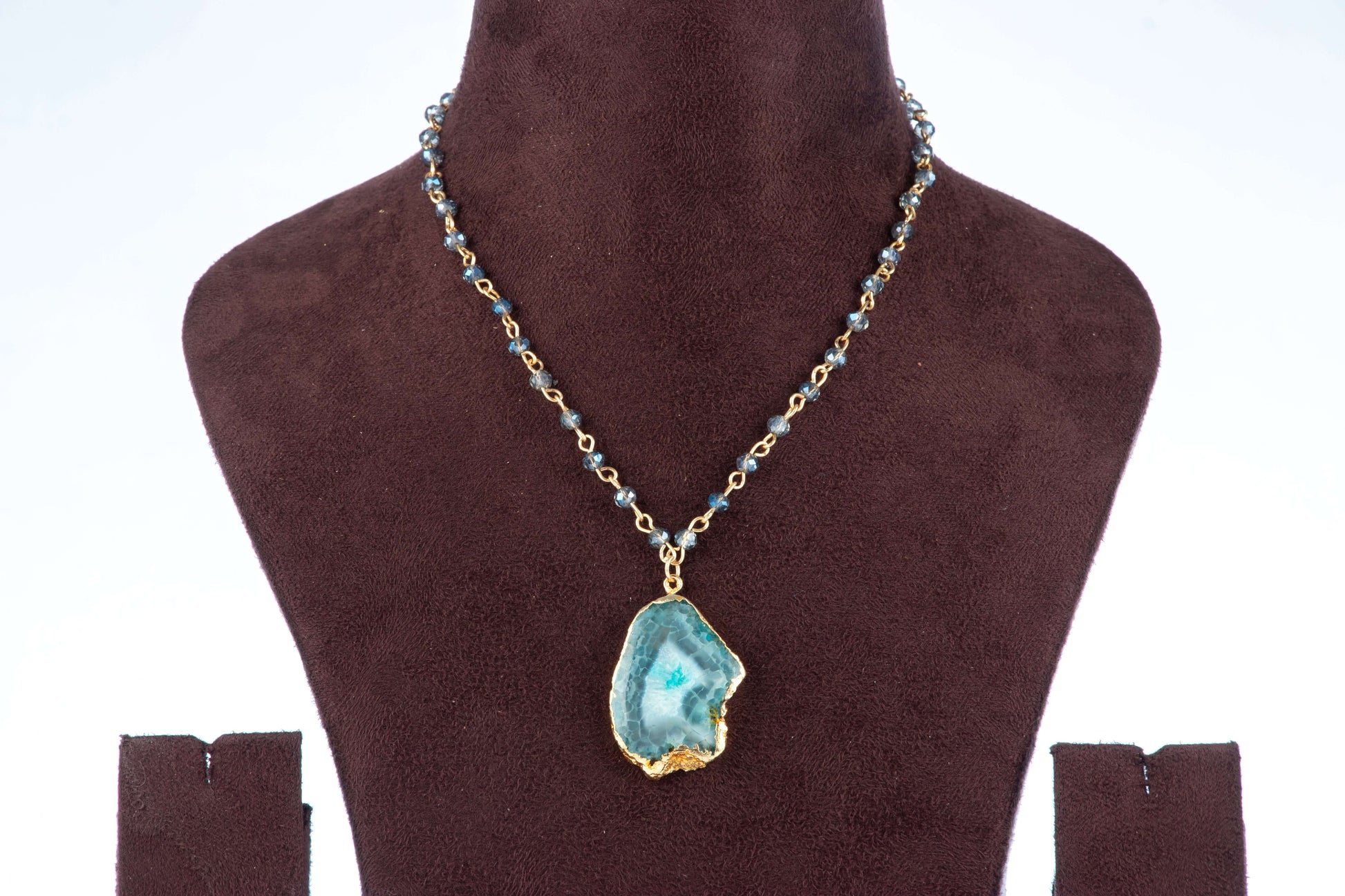 A Full beaded agate pendant necklace is shown on a brown velvet jewelry bust against a light background. The necklace features a gold-toned chain with small, faceted teal or blue beads and a large, gold-edged agate pendant with a blue-green, crystalline center.