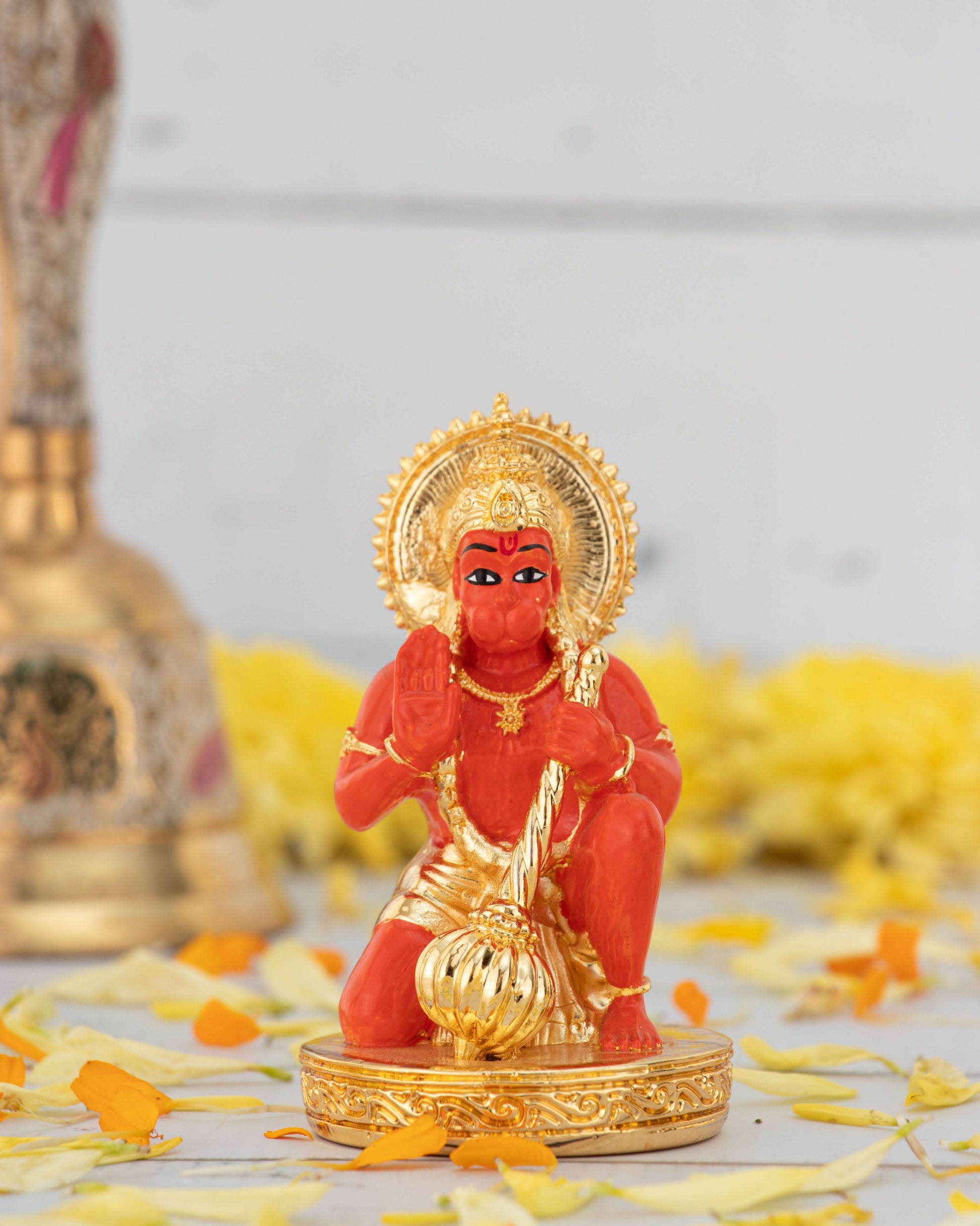 A vibrant orange-red Lord Hanuman Murti is shown kneeling on a golden, ornate base. The deity is holding a golden mace (gada) in one hand and raising the other hand in a gesture of blessing. The murti wears a golden crown and ornaments and is surrounded by a golden halo.