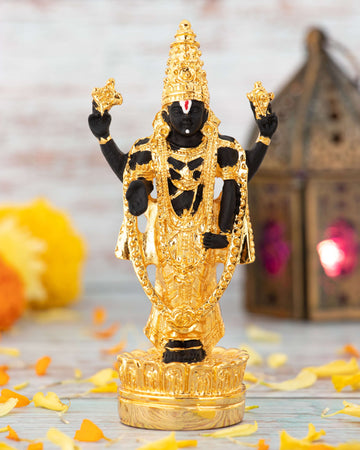 A black idol of Lord Venkateswara (Balaji) adorned with elaborate gold ornaments and a traditional white and red namam on the forehead, standing on a golden base with scattering yellow and orange marigold petals around it, against a blurred background with a lamp.