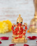 A gold-plated, 3-inch Siddhivinayak Ganesh idol with a vibrant orange or red glossy finish, seated on a golden base. The idol, a symbol of good fortune and prosperity, is shown with its trunk turned to the right. The image also features yellow flowers and red petals scattered on a light-colored wooden surface.