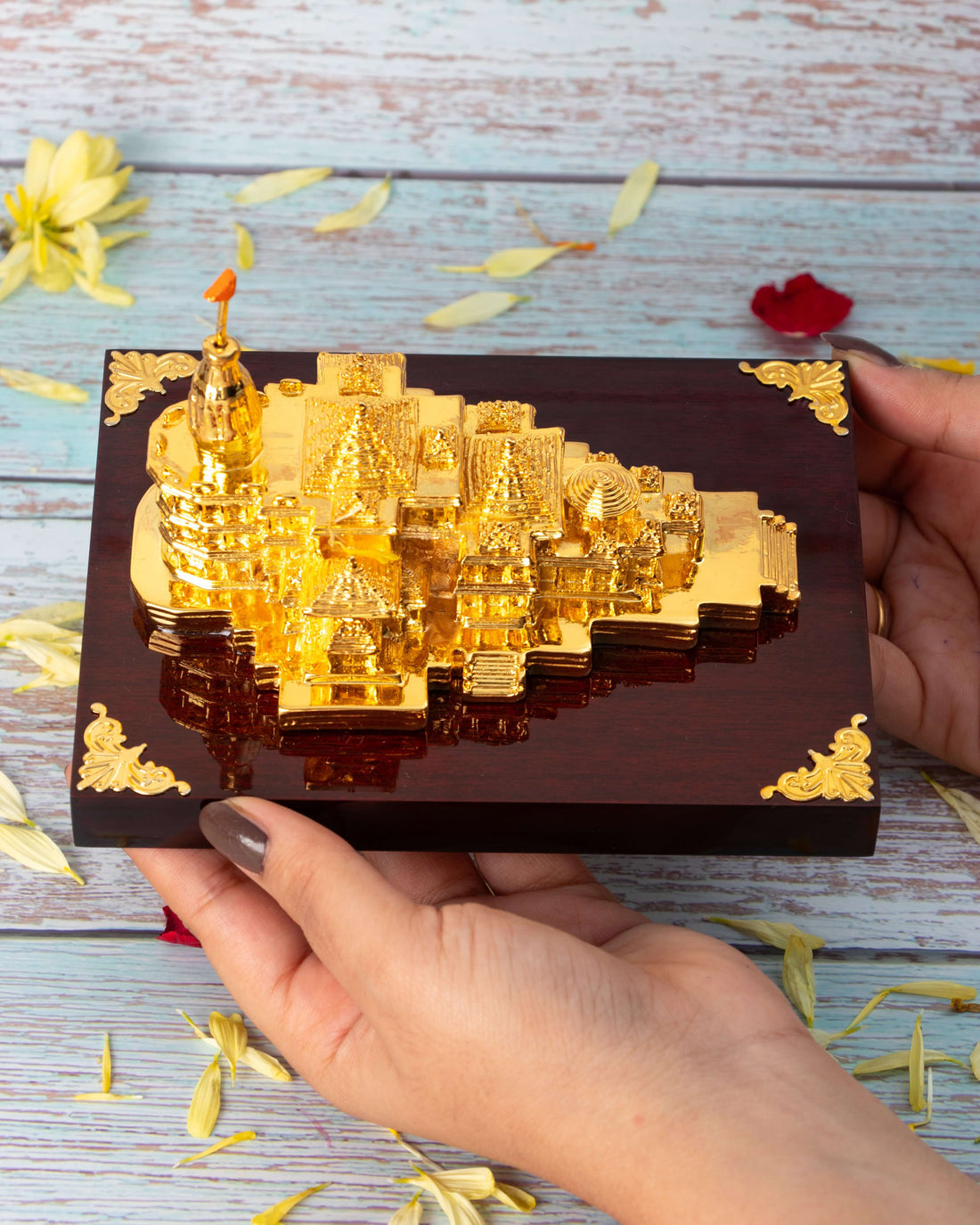 A golden, intricately detailed miniature replica of the Ram Mandir sits on a polished dark wooden base, surrounded by scattered red and yellow flower petals on a light-colored wooden surface.