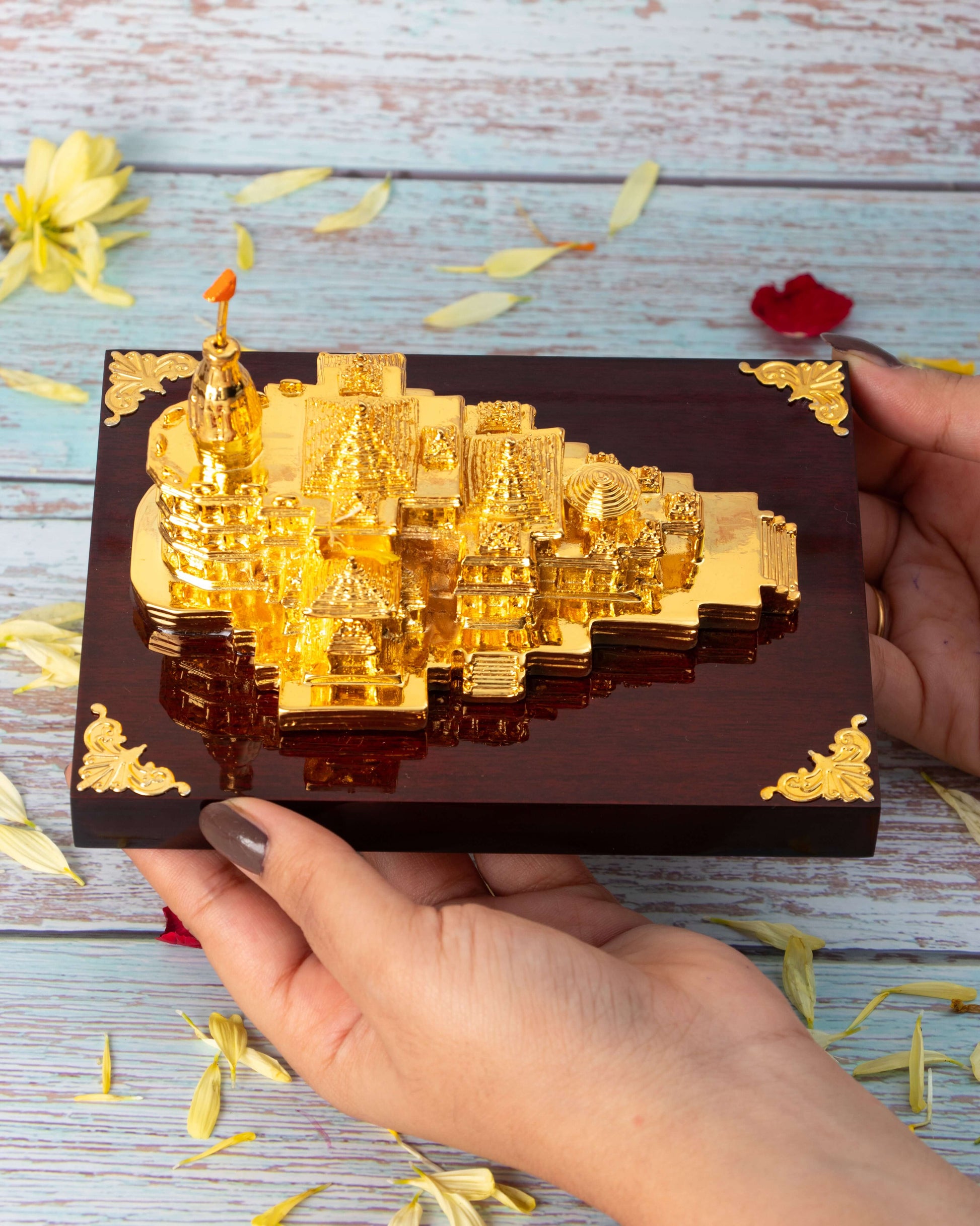 A close-up of hands holding a highly detailed, golden miniature replica of the Ram Mandir, set against a blurred, light-colored wooden background with scattered flower petals.