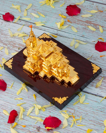 A golden, intricately detailed miniature replica of the Ram Mandir sits on a polished dark wooden base, surrounded by scattered red and yellow flower petals on a light-colored wooden surface.