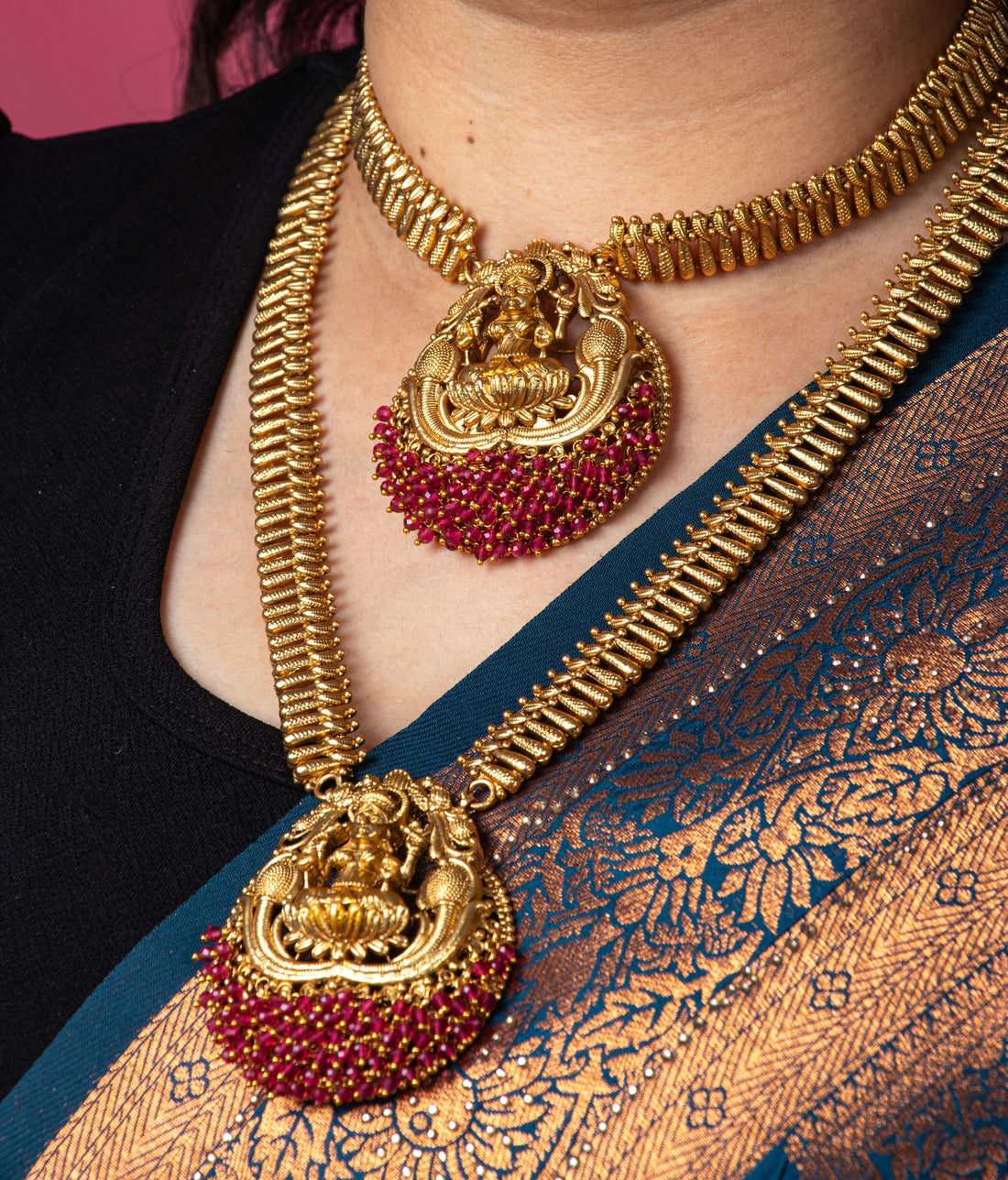 A traditional long temple necklace set with Laxmi pendants and matching earrings. The set features two gold-toned necklaces with intricate chains, both adorned with large, round pendants that depict the goddess Laxmi. The pendants are decorated with ruby-red stones and have a red beaded fringe. The matching earrings are also round with a Laxmi motif and red stone accents. The jewelry is displayed on a black bust against a white background.