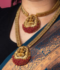 A person is wearing a traditional long temple necklace set with two gold-toned necklaces. The set includes a choker and a longer necklace, both with textured, ribbed chains. Each necklace features a large, ornate pendant depicting the goddess Laxmi, seated on a lotus. The lower part of each pendant is adorned with a cluster of ruby-red beads.