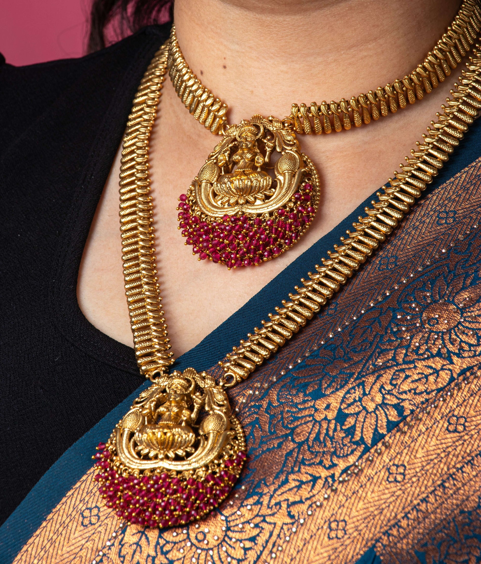 A person is wearing a traditional long temple necklace set with two gold-toned necklaces. The set includes a choker and a longer necklace, both with textured, ribbed chains. Each necklace features a large, ornate pendant depicting the goddess Laxmi, seated on a lotus. The lower part of each pendant is adorned with a cluster of ruby-red beads.
