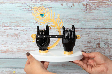 A decorative idol featuring two black hands of Lord Krishna holding a black flute, adorned with a golden peacock feather with a blue jewel and resting on a white and gold base. The idol is held by a person with manicured hands, against a blurred background of light-colored wooden planks.