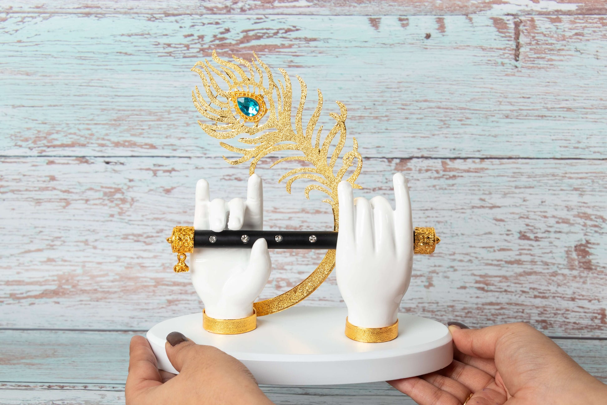 A decorative white showpiece featuring Lord Krishna's hands holding a black flute with golden details, with a large golden peacock feather adorned with a blue gem rising behind it, all set against a light blue wooden background.