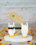 An idol depicting Lord Krishna's hands in white, holding a black flute with golden accents, and a prominent golden peacock feather with a blue gem at its eye, all resting on a white base amidst scattered orange and red flower petals, set against a light blue wooden background.